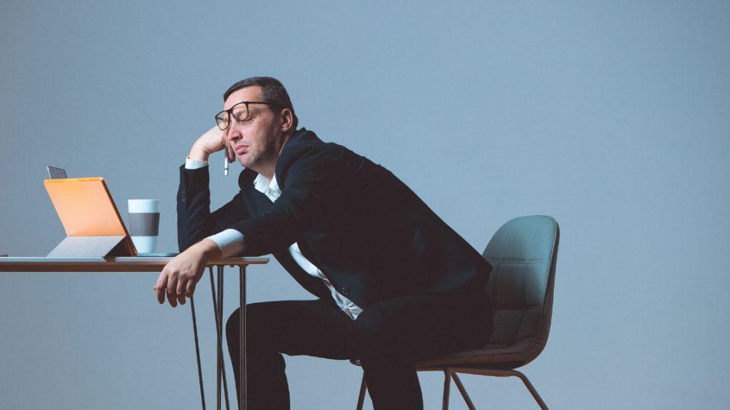 A man in a suit sits slumped over a desk. On the desk sits an open laptop.